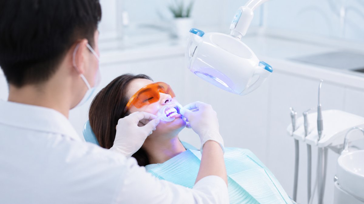 Bác sĩ nha khoa tẩy trắng răng cho khách hàng trên ghế nha khoa với màng chắn bảo vệ nướu và đèn kích hoạt